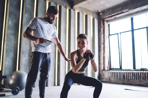 Happy strong young female in activewear squatting in gym supervised by bearded male instructor with tablet in hand and smiling at camera