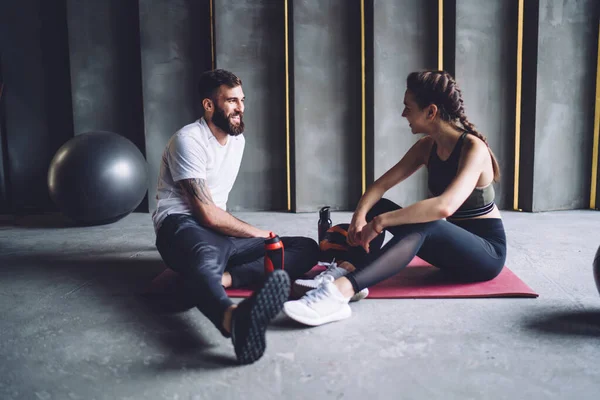 Full body of content sportive woman and man in sportswear sitting on mat and resting after workout while talking and looking at each other