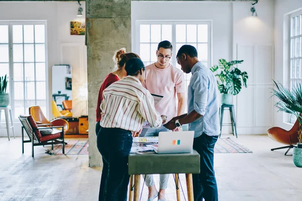 Çok kültürlü insanlar, modern ofis içlerinde bir masada duran kağıt taslaklarını analiz ediyorlar. Yetenekli hipster öğrencileri, ortak beyin fırtınası sırasında web fikirlerini tartışarak ortak iş birliği yapıyorlar.