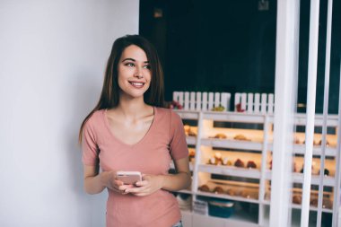 Pembe günlük giysiler içinde, beyaz duvarın yanında dikilip gülümseyen ve fırındaki bulanık vitrinlere karşı akıllı telefonla etkileşim kuran muhteşem, dalgın, koyu renk saçlı kadın.