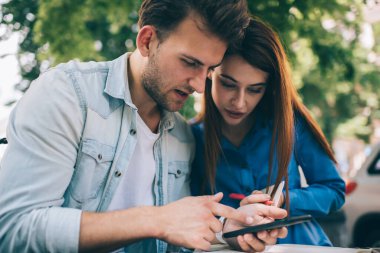 Serbest çalışan, günlük kıyafetlerle odaklanmış bir adam. Dikkatli bir bayan, güneş ışığı altında açık kafede projesini telefonda gösteriyor.