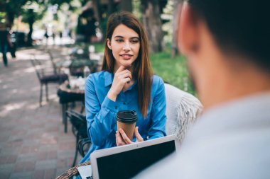 Yarı güler yüzlü kadın, masada dizüstü bilgisayarla oturmuş arkadaşını dinlerken bir fincan kahve içiyor. 