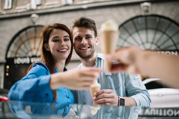 Attractive adult delighted woman in casual clothes with handsome gladful boyfriend buying cold ice cream cones while having date in downtown