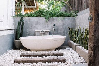 Bali 'deki lüks villanın terasında yeşil egzotik bitkiler ve çakıl taşlarıyla dekore edilmiş oval beyaz seramik küvetli modern açık hava tuvaleti.