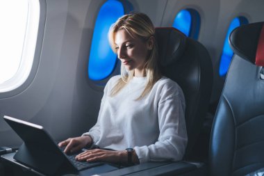 Genç, sarışın, serbest giyinen, serbest giyinen, dizüstü bilgisayarlı uçak kabini penceresinin yanındaki rahat koltukta rahatlayan bir kadın.