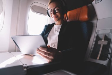 Uçak uçuşu sırasında halka açık internet sitesinde göz koruma ağı haberleri sunan optik gözlüklerde başarılı bir kadın, gürültü iptali için elektronik kulaklıklı mutlu kadın.