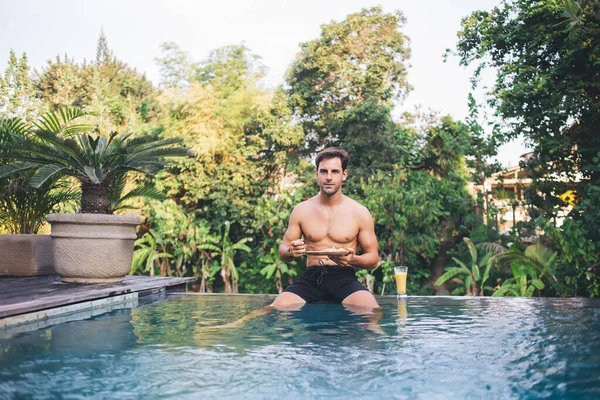 Young handsome man with naked torso in black swimming shorts sitting on edge of swimming pool and enjoying meal among vivid greenery of Bali spa resort