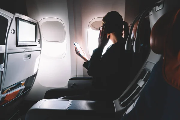 Side view of female in formal outfit listening to songs via smartphone while sitting in passenger seat near window in business class of airplane