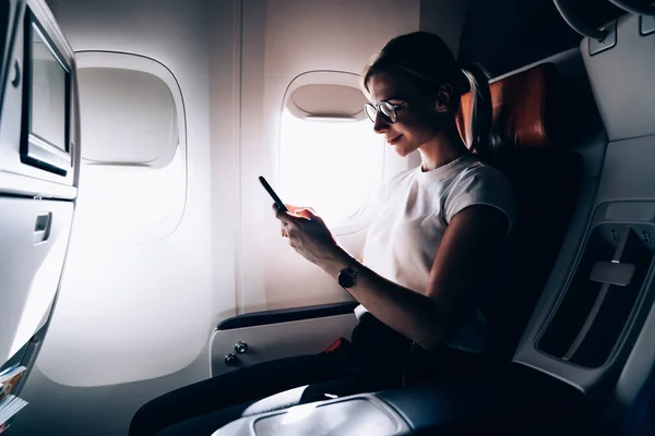 Caucasian woman passenger enjoying flight in first class with high speed wifi connection on board using smartphone and browsing internet, young female in seat near window using cellphone application