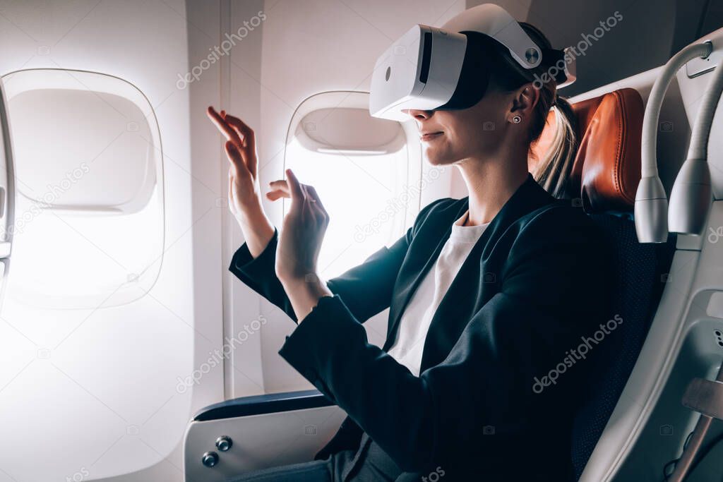 Side view of female in formal wear entertaining with virtual reality goggles and exploring air with hands while sitting in cabin of aircraft