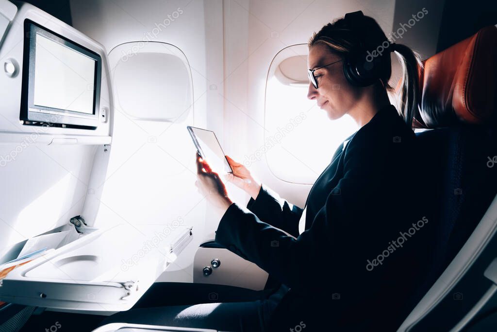 Side view of content female entrepreneur in headphones and eyeglasses enjoying videos on tablet while relaxing in passenger seat in business class of airplane