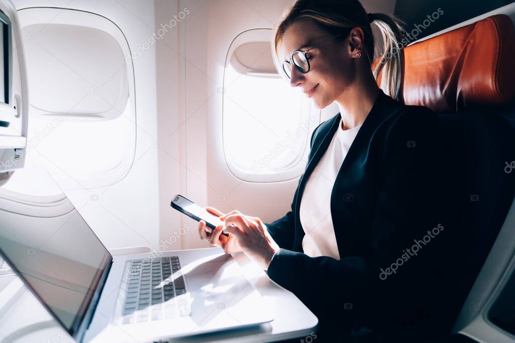 Low angle of happy thoughtful crop executive woman in glasses and elegant jacket focusing on screen and interacting with smartphone while sitting in armchair at netbook in airplane