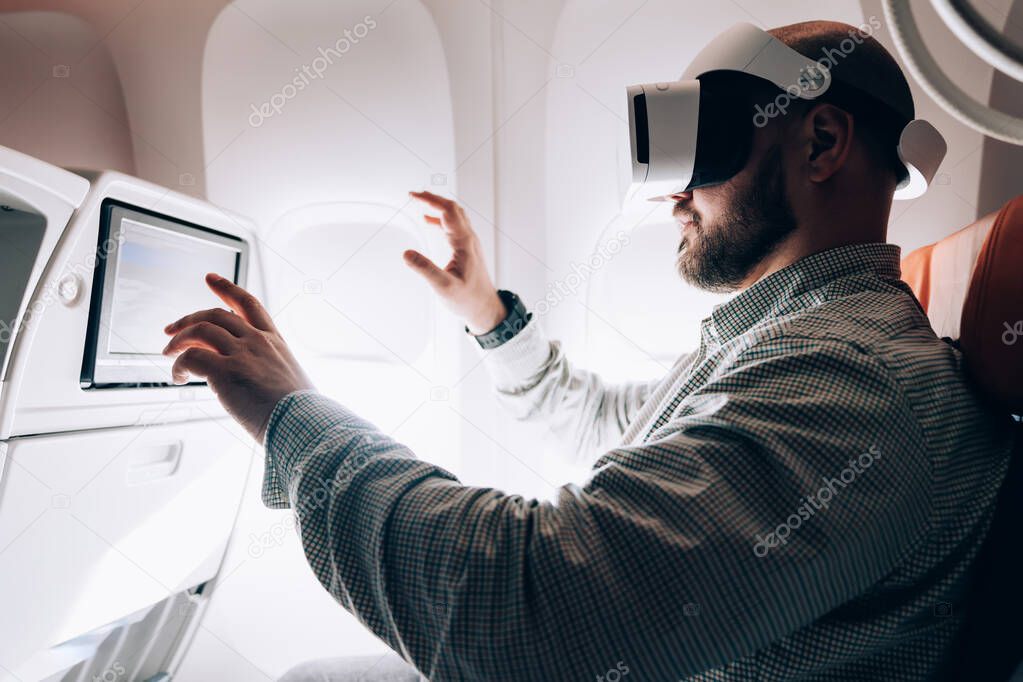 Side view of male in casual shirt sitting in VR headset in passenger seat and gesticulating while interacting with virtual reality in airplane during flight