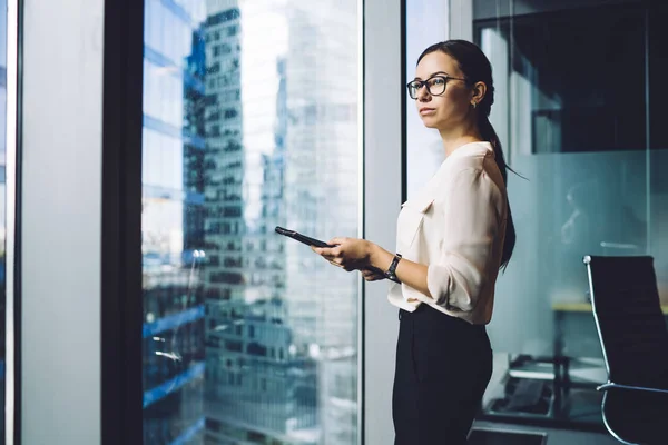 Pensive businesswoman dressed in formal apparel standing near panoramic window in enterprise company and thinking about trade investment,thoughtful female entrepreneur in eyewear for vision protection