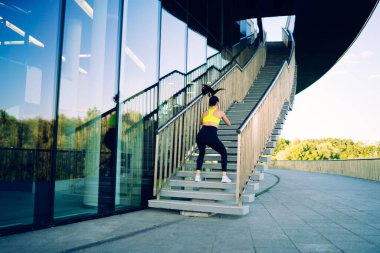 Antrenmandaki güçlü atletik kızın arkadan bakışları sıçrıyor ve dışarıda çömeliyor. Fiziksel olarak iyi durumda, beyaz bir kadın spor yapıyor. Spor yapmak için merdivenlerde canlılık ve esneklik pratiği yapıyor.