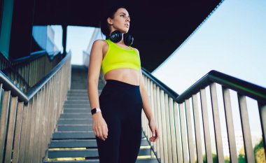 Spor kıyafetleri içindeki düşük odaklı kadın ve güneşli bir günde modern binanın merdivenlerinde duran kulaklıklarla.