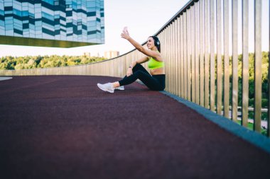 Tarz sahibi spor giyim ve beyaz spor ayakkabılı aktif genç bayan siyah kulaklıklarla bariyerin yanında otururken ve dışarıda antrenman yaparken akıllı telefon kullanırken yan görünüm