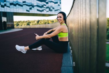 Gülümseyen genç bayan koşucunun normal spor kıyafetleri ve spor ayakkabılarıyla bariyerin yanında oturmuş kulaklıkla müzik dinlerken ve kameraya bakarken yan görüntüsü.