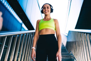 Düşük açılı spor kıyafetleri ve modern binanın merdivenlerinde duran akıllı kulaklıklı kadın koşucu.