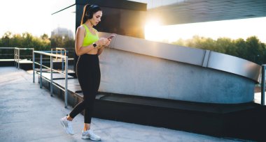 Spor kıyafetleri ve kulaklık takan, müzik dinleyen ve sosyal medyayı arka tarafta yürürken gören genç bir bayan.