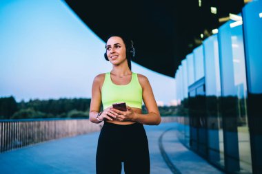 Şehirdeki cam binaların yanında duran ve cep telefonlarını tarayıp gözlerini kaçırırken kulaklıkla şarkıların tadını çıkaran modaya uygun giyinen düşük açılı bayan sporcu.