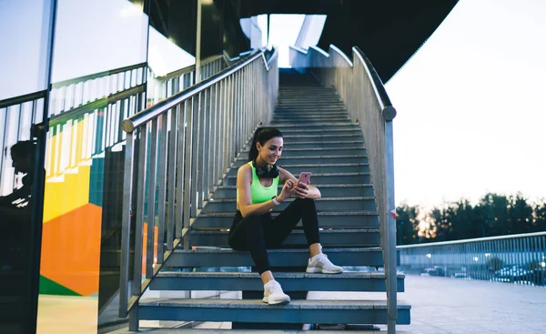 スポーツウェアやヘッドフォンの女性の笑顔の低角度携帯電話の階段に座って 夏の朝のアクティブなトレーニングの後に休息を取っている間 メッセージを読んで — ストック写真