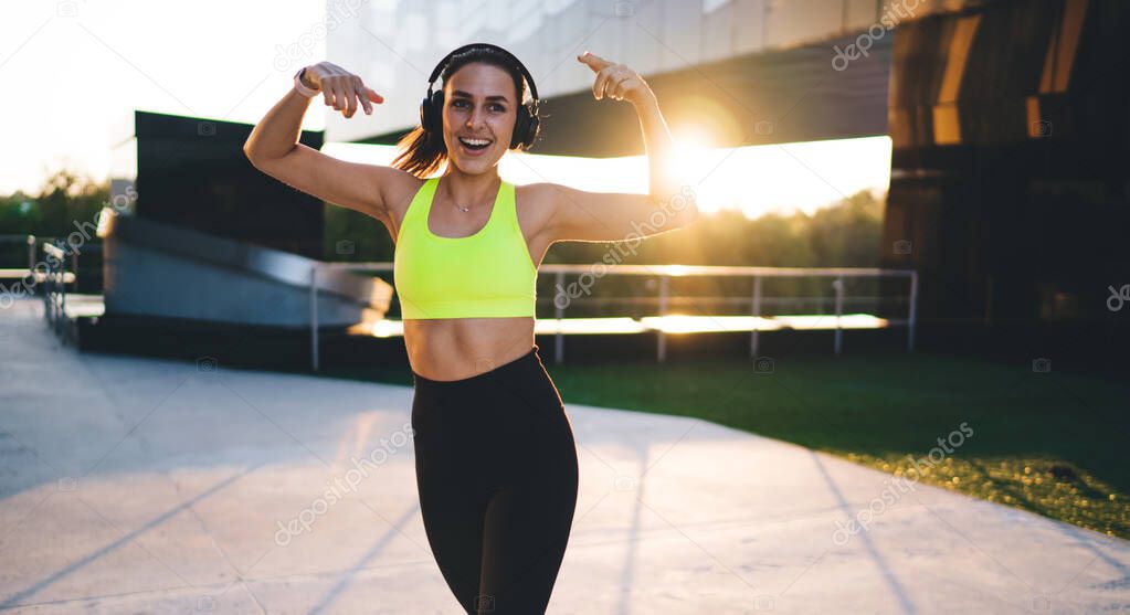 Cheerful carefree female jogger enjoying healthy lifestyle and dancing outdoors while listening positive music playlist, emotional sportive woman in bluetooth headphones listening funny podcast