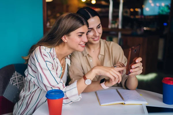 Happy female friends checking smartphone messages during coffee meeting for organisation planning in cafe interior, cheerful hipster girls browsing social networks via cellular application