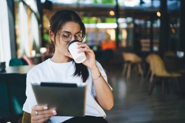 Genç bir bayan, gözlüklü, tabletli rahat bir kafede oturmuş kağıt bardaktan kahve içiyor.
