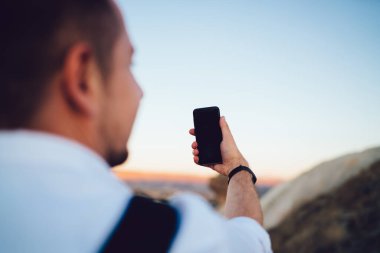 Tanımlanamayan siyah saçlı, beyaz elbiseli, açık havada cep telefonuyla selfie çekmek için poz veren erkek.