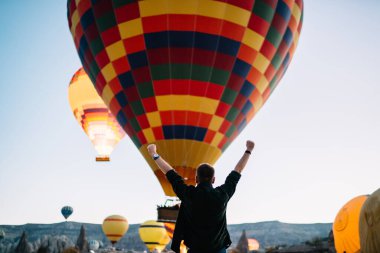 Siyah kıyafetli başarılı adamın arkası. Elleri havada kayalık dağ arazisinde uçan parlak sıcak hava balonlarını seyrediyor.