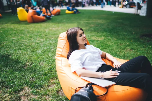 身穿休闲装的年轻女性闭着眼睛躺在橙色豆袋椅子上 通过耳机在公园里听音乐的侧影 — 图库照片