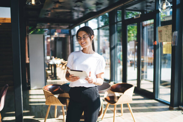 Cheerful young restaurant hostess in casual outfit and eyewear looking at camera while standing against blurred cozy interior with tablet and smiling