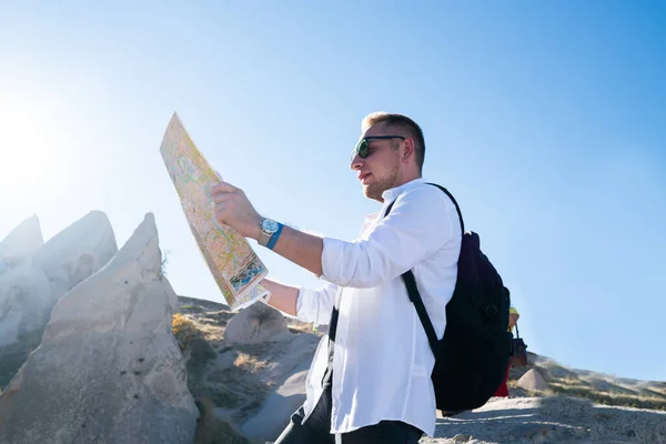 From below side view of adult man in sunglasses and with backpack using and looking at map while standing in mountain area at sunlight