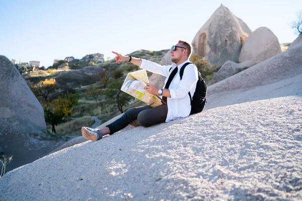 带着背包 戴着太阳镜 坐在岩石上 一边指指点点一边欣赏迷人风景的现代男性游客的侧视图 — 图库照片