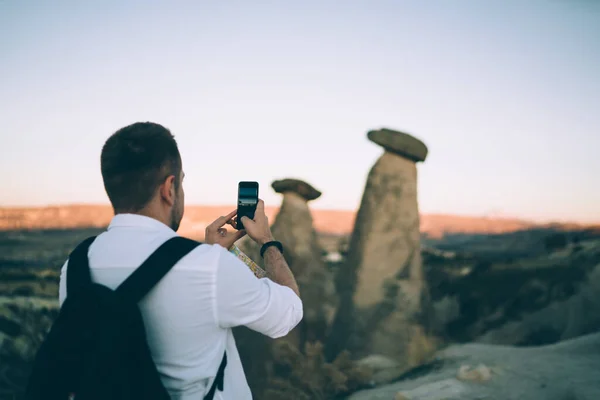 穿着白色衬衫 背着背包 用手机拍摄远处自然景观的无法辨认的棕色头发男性 — 图库照片