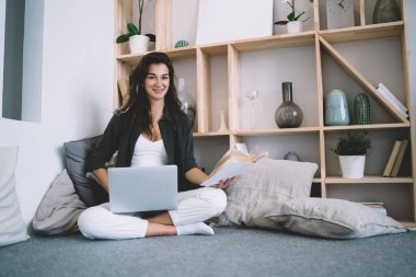 Kanepede oturup dizüstü bilgisayarda çalışan, elinde kitap tutan ve kameraya bakan serbest çalışan genç bir kadın.