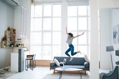 Karantina sırasında evde eğlenirken rahat koltukta zıplayıp siyah saçlı, mutlu, ince bir kadın vücudu.