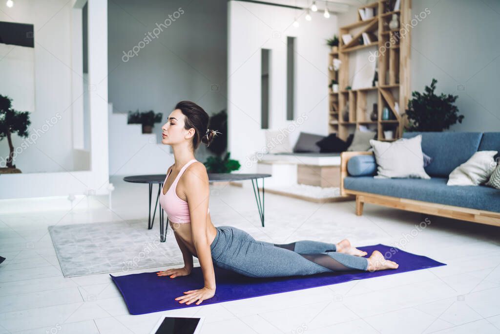 Side view full length of slim female with eyes closed in pink top and grey leggings doing sphinx pose on mat at modern living room at home