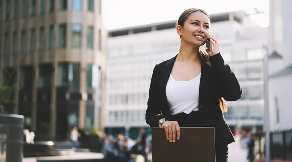 スマートフォンで話し ノートパッドを手にしながら通りに立って離れて見ている正式な摩耗で陽気な若いビジネスマンの女性 — ストック写真