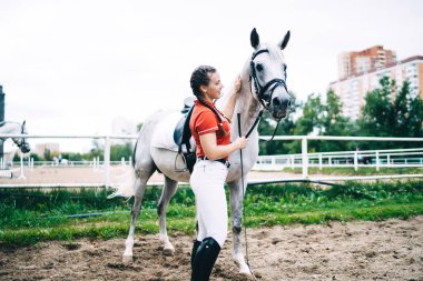 Sıradan tişörtler ve jokey çizmeleri giyen, otlakta durup hayvanlara nazikçe dokunan, safkan atları okşayan profesyonel kadın binicinin yan görüntüsü.