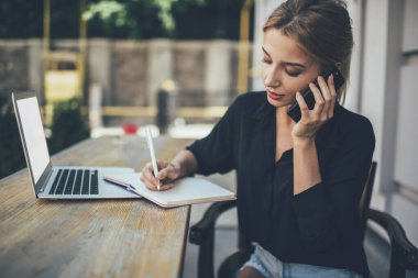 Eğitimle ilgili ders kitabında bilgi toplayan yetenekli kız öğrenci dışarıda dizüstü bilgisayarla cep telefonuyla eğitim alırken, akıllı telefon aracılığıyla serbest çalışan öğrenci iletişim kuruyor.