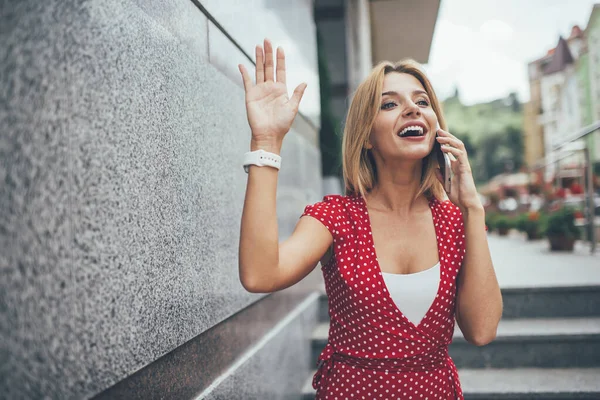 Trendy dressed woman with cute smile on face using cellphone gadget for making roaming phoning and talking about meeting time, happy girl waving at street enjoying friendly mobile conversation