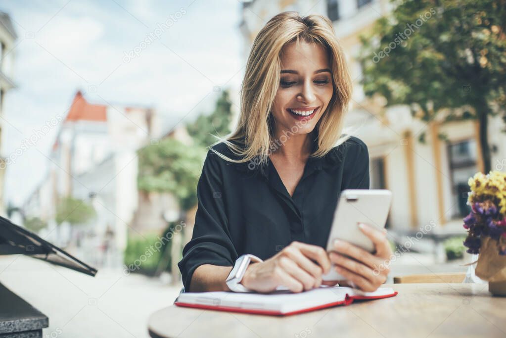 Cheerful female blogger using textbook for planning publication posting sitting in sidewalk and reading notification from social networks, happy hipster girl watching streams video on cellphone