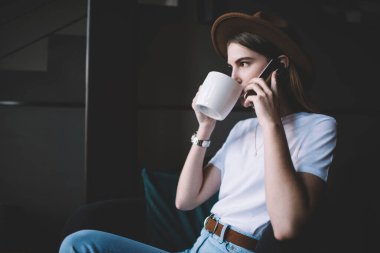 Şapkalı, beyaz bardaktan sıcak içecek içen ve kafede rahat bir koltukta otururken akıllı telefondan konuşan genç bir kadının yan görüntüsü.