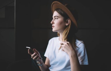 Moda şapkalı, şık giyinmiş, akıllı telefonu elinde tutarken yüzünü çeviren genç ve düşünceli bir hanımın yan görüntüsü.