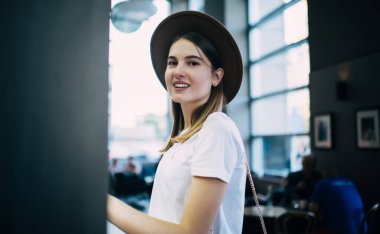 Rahat bir kafede dikilip hafta sonu dinlenirken kameraya bakan rahat tişörtlü, gülümseyen genç bir kadının yan görüntüsü.