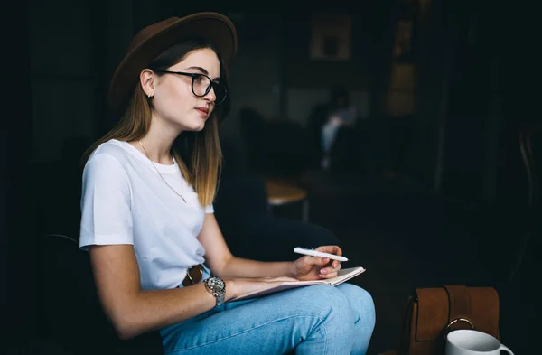 Pensivo Jovem Blogueira Óculos Chapéu Sentado Interior Café Fazendo Anotações — Fotografia de Stock