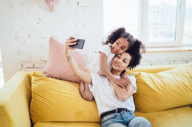 Siyah tatmin olmuş genç bir anne ve kıvırcık saçlı sevimli bir kız çocuğu sarı kanepede, yastıklı akıllı telefonla fotoğraf çekiyorlar.
