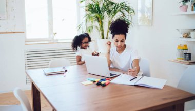 Beyaz nedensel kıyafetli ve kıvırcık saçlı genç etnik anneden. Modern evde dizüstü bilgisayar kullanarak çalışırken küçük kızıyla birlikte masa başında notlar yazıyorlar.